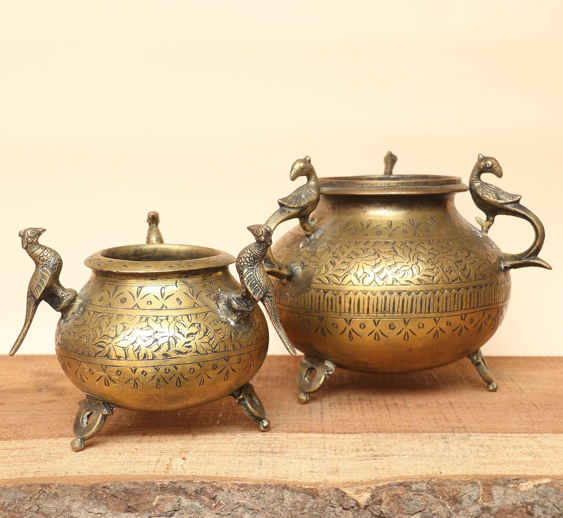 A set of traditional Indian brass home decor items, including a large ornate pitcher with an elephant head spout, a small brass teapot, a dark brass pot, and a brass elephant figurine, arranged on a wooden shelf against a warm beige background.
