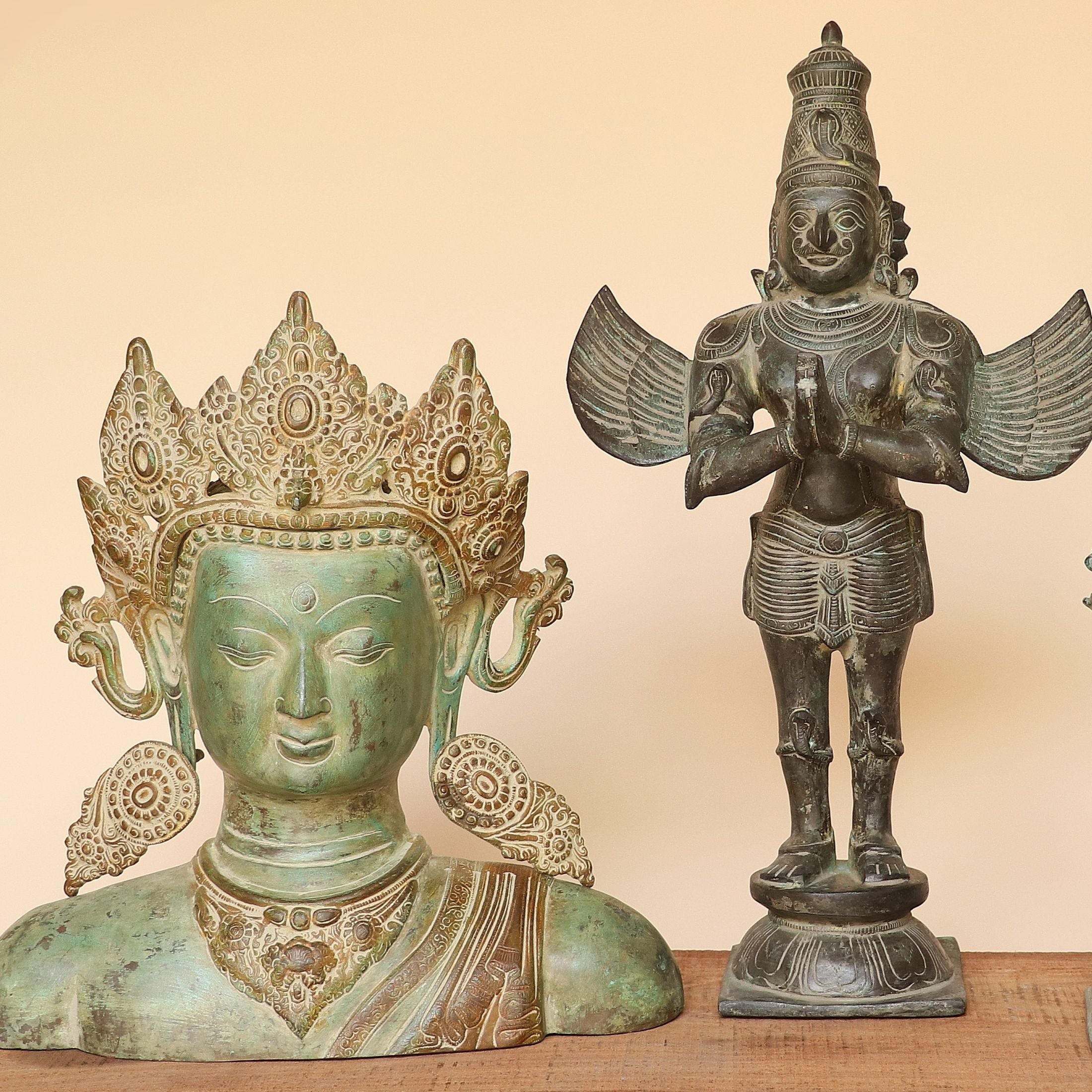 Three decorative metal statues with a greenish patina are displayed on a wooden surface against a plain beige wall. The statues include a large crowned face bust, a standing winged figure, and a smaller dancing deity figure.