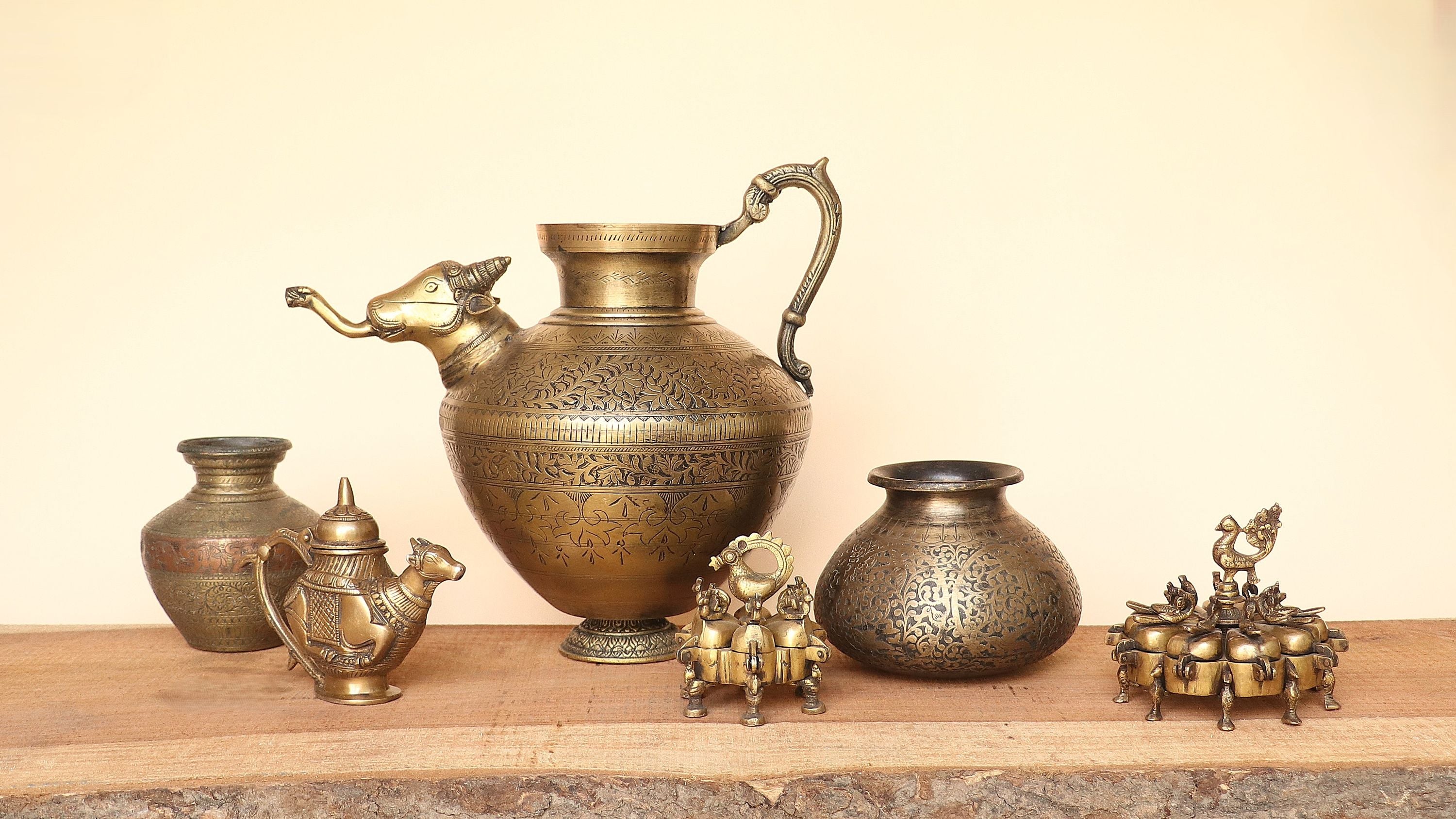 A collection of antique brass and bronze decorative metal vessels, including a large ewer, a camel-shaped pitcher, a small pot, a round jar, and ornate figurines, arranged on a wooden shelf against a plain beige background.
