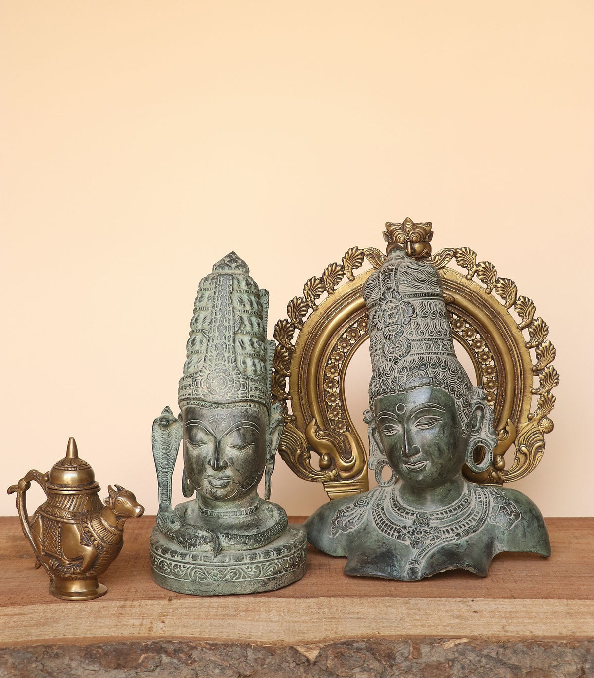Two ornate bronze deity bust statues with detailed headdresses and jewelry, displayed on a wooden surface against a plain beige background, alongside a small brass pot.