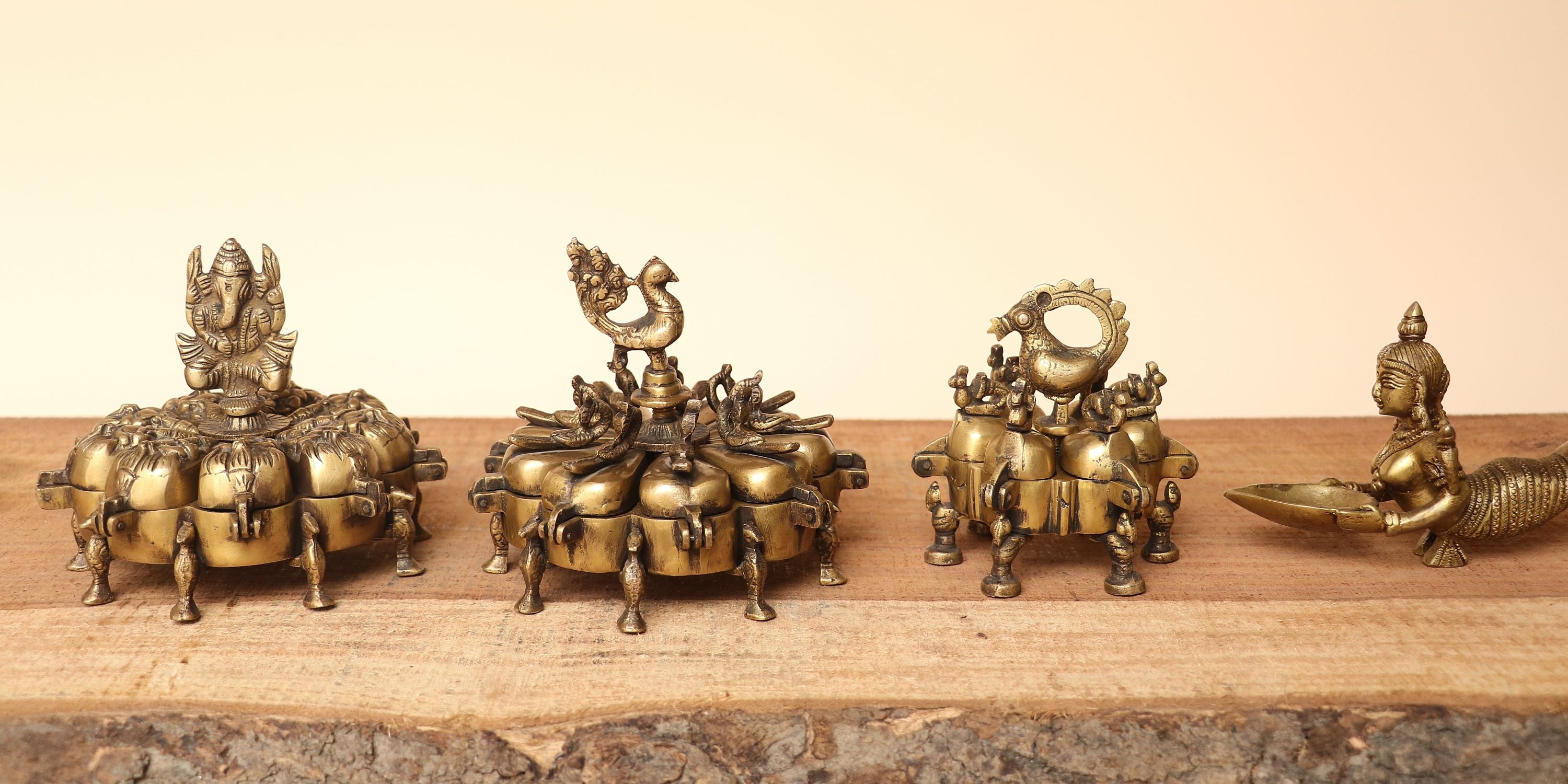 Four ornate brass decorative figurines arranged in a row on a rustic wooden surface against a plain beige background. The sculptures appear to depict mythological or traditional figures, with detailed metallic finishes and varied shapes.