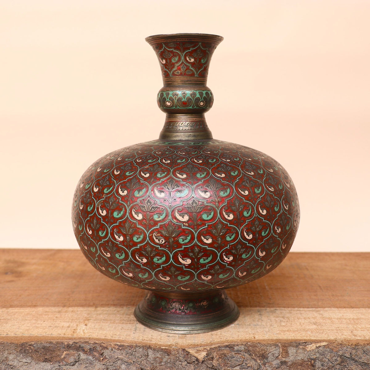 An antique-style decorative vase with a bulbous body, narrow neck, and pedestal base, decorated with an intricate repeating floral pattern in dark red, green, and gold tones, displayed on a wooden surface against a solid beige background.